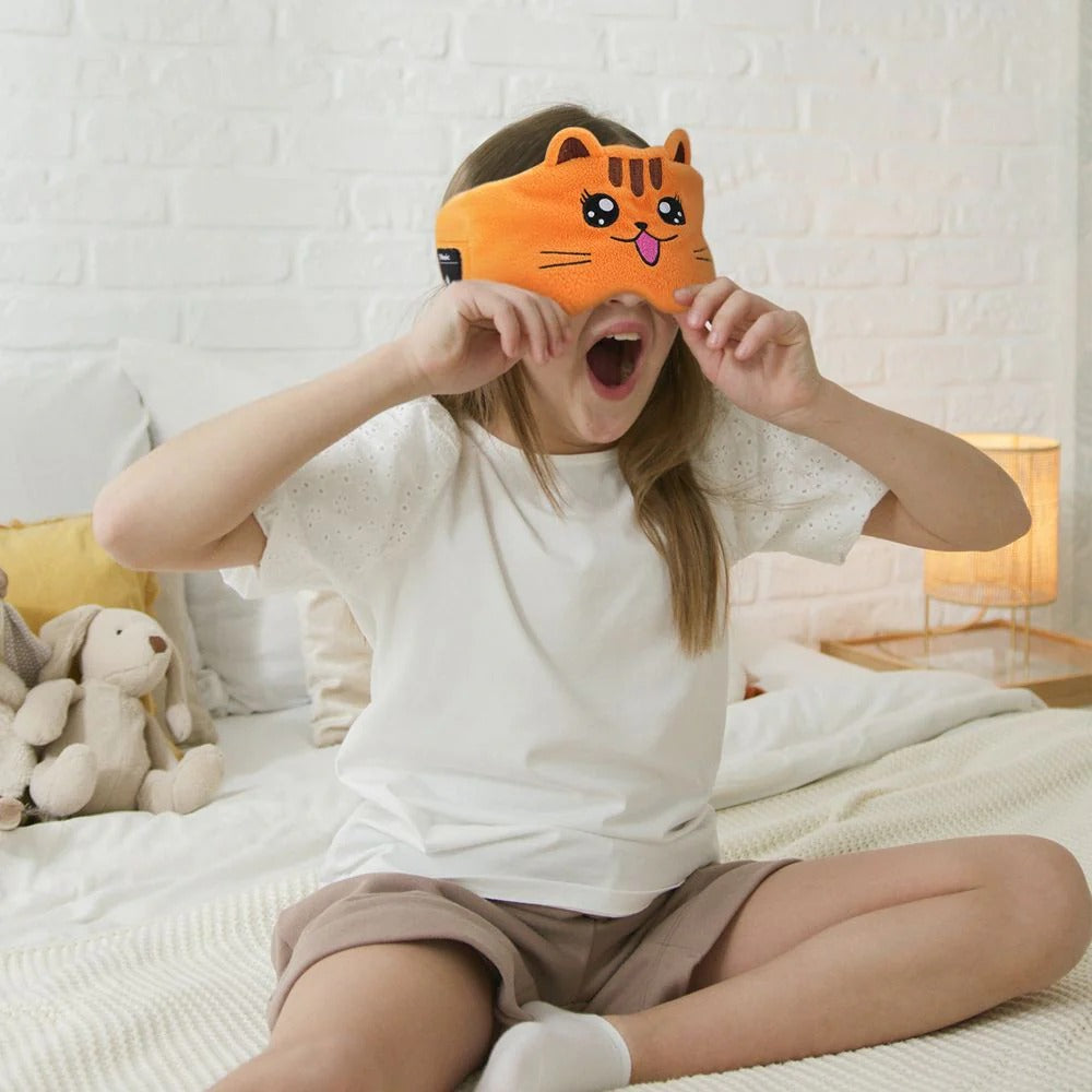 Person wearing a cartoon cat eye mask on a bed with a white brick wall background. JOEYZ_soft_headphones_tiger_sleeping