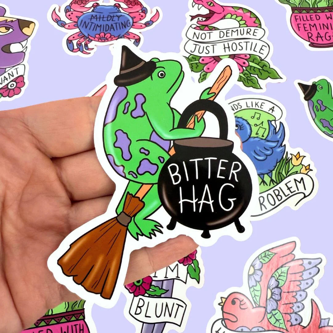 Hand holding a sticker of a green frog character with a pot labeled 'Bitter Hag' against a background of other colorful stickers.