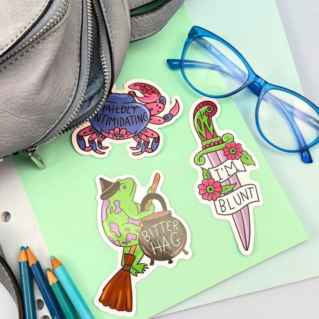 Three colorful stickers on a green surface with a bag, glasses, and pens in the background.