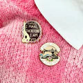 Two gold pins with engraved designs on a pink fabric background