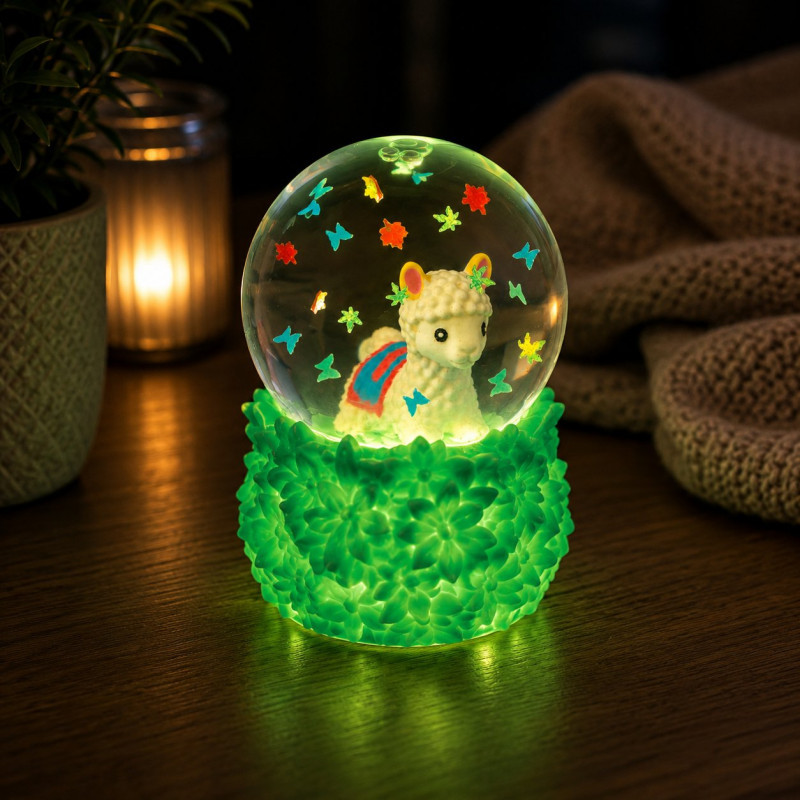 A glowing decorative snow globe with a small white llama figure inside, surrounded by colorful star and butterfly decorations, set on a green textured base on a wooden surface with warm ambient background lighting. Smooshos_alpaca_snow_dome_squishiy_glowing_in_the_dark