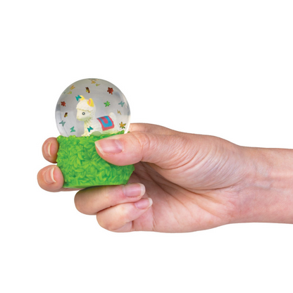 A hand holding a small stress toy snow globe with a green grass-textured base and a clear round top containing a cartoon figure and colorful star details, shown against a plain white background. Smooshos_alpaca_snow_dome_squishiy_in_hand