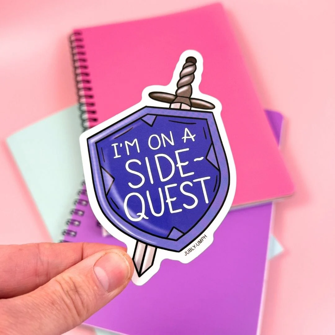 Sticker with 'I'm on a side-quest' design held by a hand against a colorful background. Sticker_Jubly-umph_sidequest_sticker_book