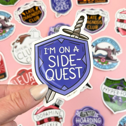 Sticker with 'I'm on a side-quest' design held over a pink background with various other stickers. Sticker_Jubly-umph_sidequest_sticker_sticker_background
