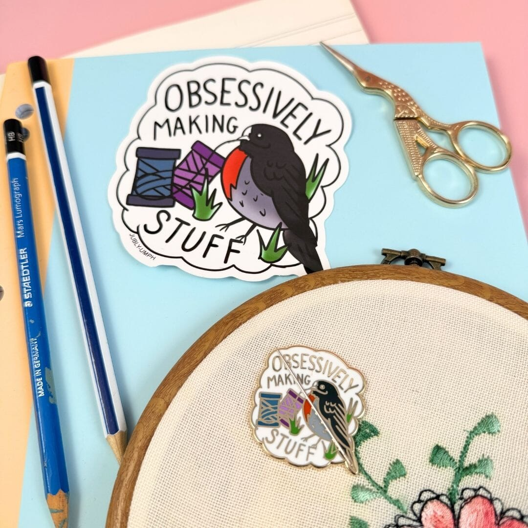 Sticker with a bird design and text 'Obsessively Making Stuff' on a blue background with embroidery hoop and tools. Sticker_Jubly_Umph__Obsessively_making_stuff_crafting